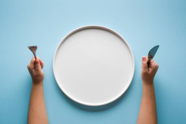 Çatal, bıçak ve mavi arka planda boş tabak tutan kızların üst görüntüsü.