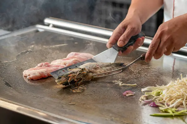 Şef sıcak teppanyaki ızgara yüzeyinde taze karides pişiriyor.