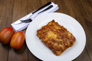Bolonez soslu lazanya, jambon peyniri, domates sosu ve kıyma. Beyaz tabakta servis edilen yemekler. Peçetedeki çatal bıçak takımı..