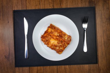 Bolonez soslu lazanya, jambon peyniri, domates sosu ve kıyma. Sağında çatal, solunda bıçak olan beyaz bir tabakta servis ediliyor..