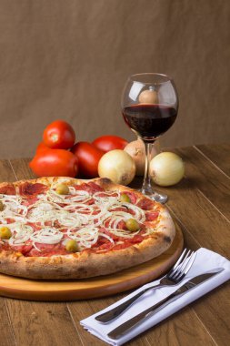 Sucuklu pizza, soğan dilimleri, mozzarella peyniri ve yeşil zeytinli gurme fotoğrafı. Bir kadeh kırmızı şarap. Görüntüyü oluşturmak için kırsal formundaki bileşenler.