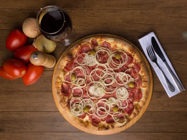 Pepperoni pizza with slices of onions, mozzarella cheese and green olives. Glass of wine, onions and rustic tomatoes to compose the photo. Top gastronomic photography.