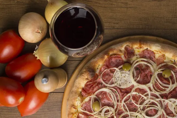 Pepperoni pizza with slices of onions, mozzarella cheese and green olives. Glass of wine, onions and rustic tomatoes to compose the photo. Gastronomic close-up photograph of Topo.