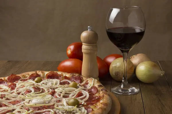 Pepperoni pizza with slices of onions, mozzarella cheese and green olives. Glass of wine, onions and rustic tomatoes. Horizontal Gastronomic Photography with upper left space for texts.