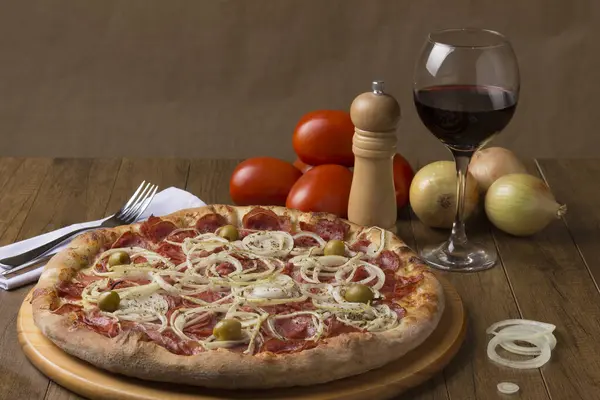 Pepperoni pizza with slices of onions, and green olives. Glass of red wine, onions and rustic tomatoes. Vertical Gastronomic Photography with upper left space for texts.