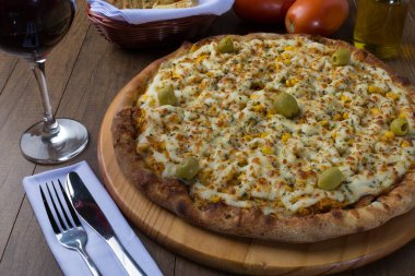 Tahta bir tahtanın üzerinde servis edilen lezzetli tavuk aromalı pizza. Mozarella, tavuk, krem peynir, yeşil zeytin, mısır ve kırmızı domates sosuyla yapılmış. Bir bardak kırmızı şarap, crostini aperatifleri, beyaz peçeteye çatal ve bıçak. Yatay Fotoğrafçılık