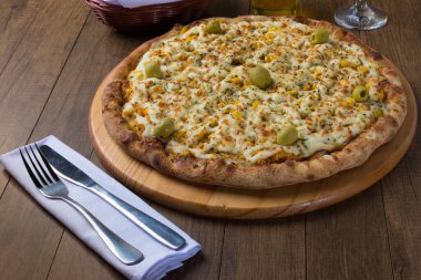 Tahta bir tahtanın üzerinde servis edilen lezzetli tavuk aromalı pizza. Mozarella, tavuk, krem peynir, yeşil zeytin, mısır ve kırmızı domates sosuyla yapılmış. Çatal ve bıçak beyaz peçeteye. Yatay Fotoğrafçılık.