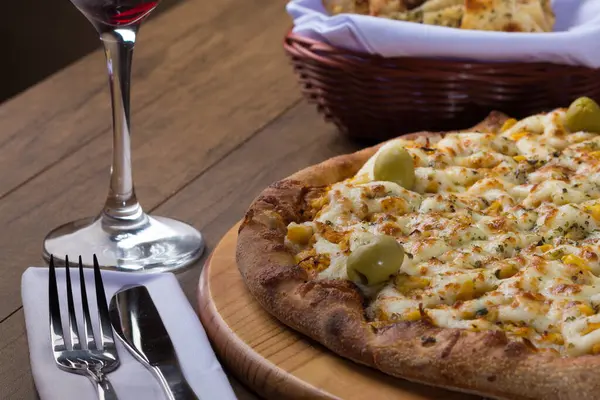Ahşap bir tahtanın üzerinde servis edilen Catupiry Chicken aromalı pizza. Mozarella, tavuk, krem peynir, yeşil zeytin, mısır ve kırmızı domates sosu. Bir bardak kırmızı şarap ve crostini. Yatay Fotoğrafçılık.
