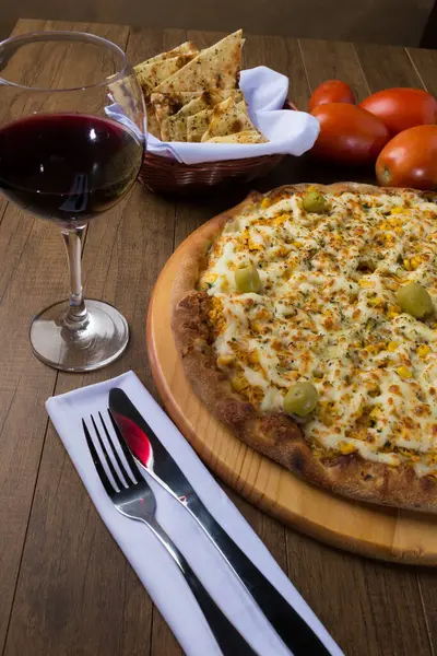 Tahta bir tahtanın üzerinde servis edilen lezzetli tavuk aromalı pizza. Mozarella, tavuk, krem peynir, yeşil zeytin, mısır ve kırmızı domates sosuyla yapılmış. Bir bardak kırmızı şarap, crostini aperatifleri, beyaz peçeteye çatal ve bıçak. Dikey fotoğrafçılık.