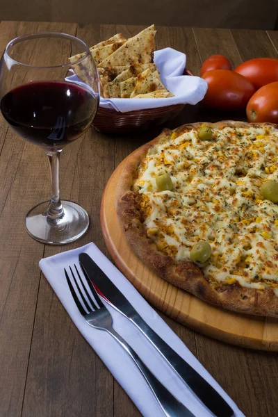 Tahta bir tahtanın üzerinde servis edilen lezzetli tavuk aromalı pizza. Mozarella, tavuk, krem peynir, yeşil zeytin, mısır ve kırmızı domates sosuyla yapılmış. Bir bardak kırmızı şarap, crostini aperatifleri.