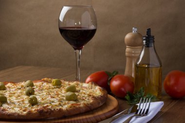 Gratin Loin Pizza. Mozzarella, Kanada usulü sığır filetosu, kremalı süt ve yeşil zeytin. Yanında bir bardak kırmızı şarap, kırmızı domates, zeytinyağı, çatal ve bıçak beyaz peçete ve maydanozda..