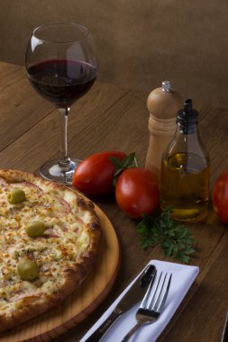 Gratin Loin Pizza. Mozzarella, Kanada usulü sığır filetosu, kremalı süt ve yeşil zeytin. Yanında bir bardak kırmızı şarap, kırmızı domates, zeytinyağı, çatal ve bıçak beyaz bir peçete ve maydanoz. Dikey.