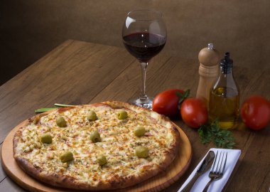 Gratin Loin Pizza. Mozzarella, Kanada usulü sığır filetosu, kremalı süt ve yeşil zeytin. Yanında bir bardak kırmızı şarap, kırmızı domates, zeytinyağı, karabiber öğütücü, çatal ve bıçak beyaz bir peçete ve maydanoz. Yatay fotoğraf.