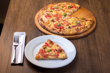 Mozzarella, peynir, domates, fesleğen ve yeşil zeytinle yapılan Marguerita aromalı pizza diliminin fotoğrafı. Geleneksel Napoli pizzası. Arka planda pizza ve solda çatal bıçak takımı var..