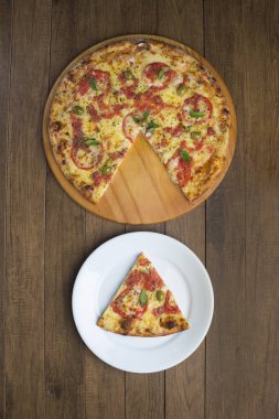 Mozzarella peyniri, domates, fesleğen yaprağı ve yeşil zeytinlerle yapılmış bir pizza fotoğrafı ahşap bir tahtada servis ediliyor. Pizza dilimi alt köşedeki pizzayla aynı hizada servis ediliyor..