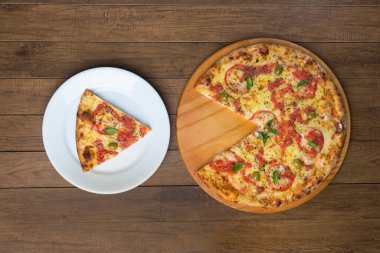 Mozzarella peynirli, domatesli, fesleğenli ve yeşil zeytinli pizza ahşap bir tahtada servis edilir. Tabakta servis edilen pizza dilimi. Tabak solda, pizza sağda. Üst resim.