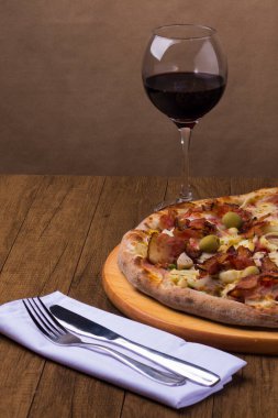 Jambon, bezelye yumurtası, palmiye, pepperoni, soğan, mozzarella ve pastırmadan yapılmış Portekiz pizzası. Bir kadeh kırmızı şarap. Beyaz bir peçeteye çatal ve bıçakla dikey gastronomik fotoğraf..