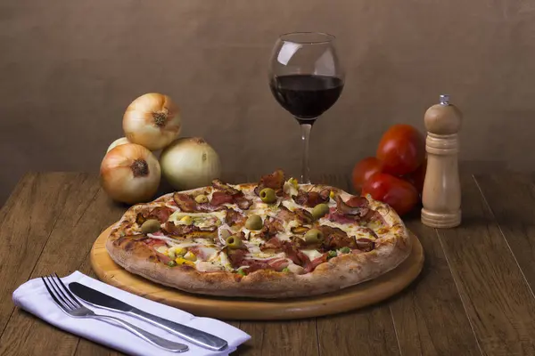 Jambon, bezelye yumurtası, palmiye, pepperoni, soğan, mozzarella ve pastırmadan yapılmış Portekiz pizzası. Yemeklere eşlik etmesi için tahta bir tahtada kırmızı şarapla servis edilmiş. Çatal ve bıçak..