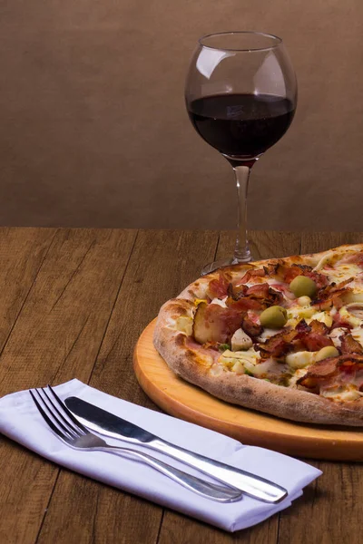 Jambon, bezelye yumurtası, palmiye, pepperoni, soğan, mozzarella ve pastırmadan yapılmış Portekiz pizzası. Bir kadeh kırmızı şarap. Beyaz bir peçeteye çatal ve bıçakla dikey gastronomik fotoğraf..