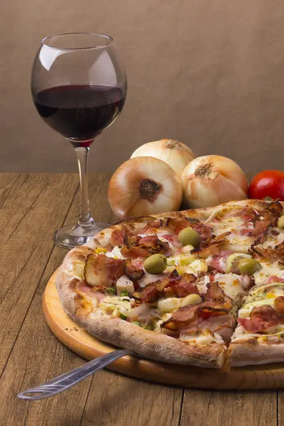 Jambon, bezelye yumurtası, palmiye, pepperoni, soğan, mozzarella ve pastırmadan yapılmış Portekiz pizzası. Bir kadeh kırmızı şarap. Pizza dilimi servis etmek için kesilmiş. Pizzacıların gurme fotoğrafı.