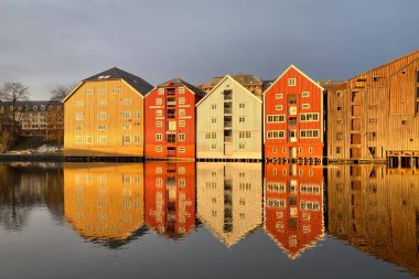 Norveç, Trondheim 'ın Brygge ilçesinde Nidelva Nehri ile birlikte kış güneşinde bulutlu gökyüzüne karşı renkli tarihi kereste depoları.