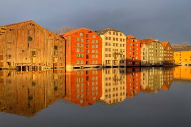 Norveç, Trondheim 'ın Brygge ilçesinde Nidelva Nehri ile birlikte kış güneşinde bulutlu gökyüzüne karşı renkli tarihi kereste depoları.