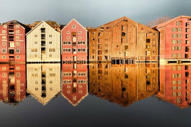 Norveç, Trondheim 'ın Brygge ilçesinde Nidelva Nehri ile birlikte kış güneşinde bulutlu gökyüzüne karşı renkli tarihi kereste depoları.