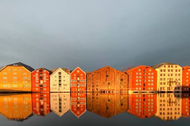 Norveç, Trondheim 'ın Brygge ilçesinde Nidelva Nehri ile birlikte kış güneşinde bulutlu gökyüzüne karşı renkli tarihi kereste depoları.