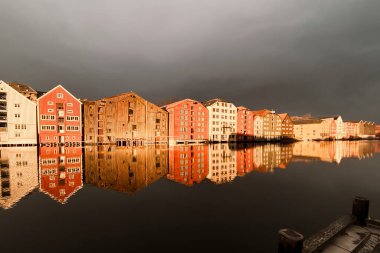 Norveç, Trondheim 'ın Brygge ilçesinde Nidelva Nehri ile birlikte kış güneşinde bulutlu gökyüzüne karşı renkli tarihi kereste depoları.