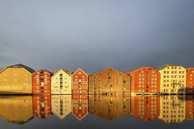 Norveç, Trondheim 'ın Brygge ilçesinde Nidelva Nehri ile birlikte kış güneşinde bulutlu gökyüzüne karşı renkli tarihi kereste depoları.