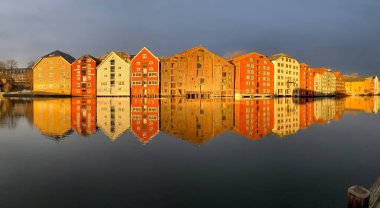 Norveç, Trondheim 'ın Brygge ilçesinde Nidelva Nehri ile birlikte kış güneşinde bulutlu gökyüzüne karşı renkli tarihi kereste depoları.