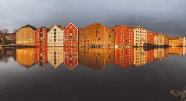 Norveç, Trondheim 'ın Brygge ilçesinde Nidelva Nehri ile birlikte kış güneşinde bulutlu gökyüzüne karşı renkli tarihi kereste depoları.