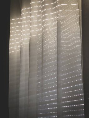 Açık renkli perdeler kapalı bir pencerenin kepenklerinden içeri giriyor.