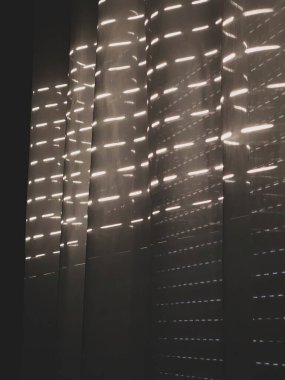 Açık renkli perdeler kapalı bir pencerenin kepenklerinden içeri giriyor.