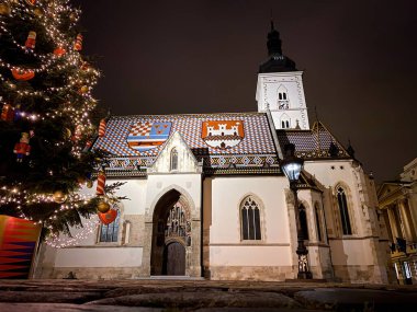 Hırvatistan 'ın Zagreb kentindeki St. Mark Kilisesi' nin ışıklandırılmış düşük açılı manzarası ve kışın gece vakti Noel ağacı