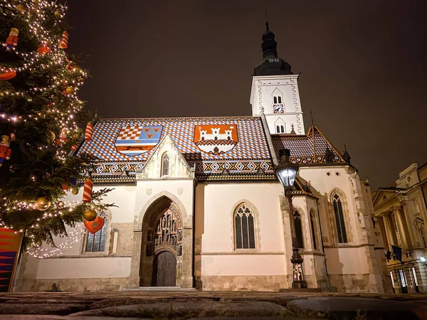 Hırvatistan 'ın Zagreb kentindeki St. Mark Kilisesi' nin ışıklandırılmış düşük açılı manzarası ve kışın gece vakti Noel ağacı