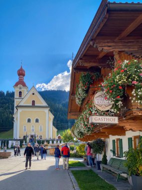 Ben Wilden Kaiser, Avusturya - 10 Ekim 2021: İnsanlar kilisede çiftlik evi ve çiçeklerle ön planda yürüyorlar - ünlü bir televizyon arkaplanı 