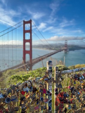 San Francisco, ABD - 12 Nisan 2024: Golden Gate Köprüsü 'nün manzaralı manzarası ve ön planda mavi gökyüzü ile bulutlu bir çitin üzerinde birçok asma kilit
