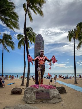 Honolulu, Hawaii, USA - April 2, 2024: Duke Paoa Kahanamoku Statue decorated with flowers at Waikiki Beach under palm trees against blue sky with clouds clipart