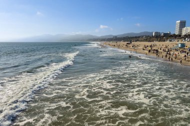 Dalgaların olduğu kıyı şeridi manzarası ve Santa Monica Sahili 'nde mavi gökyüzüne karşı plaj hayatından keyif alan insanlar.