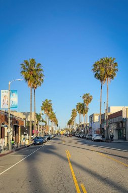 Venice Beach, Kaliforniya, ABD - 18 Nisan 2024: Venedik Plajı, Los Angeles 'taki Abbot Kinney Bulvarı' nda uzun palmiye ağaçları, yerel dükkanlar ve mavi gökyüzüne karşı yol boyunca park edilmiş arabalar