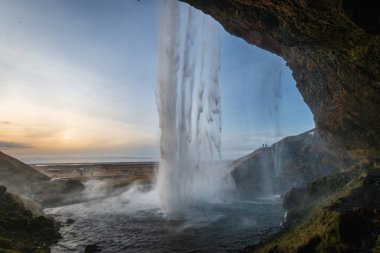 İzlanda 'daki Seljalandsfoss şelalesinin arkasından manzara, gün batımında geniş bir açık manzaranın önüne akan su