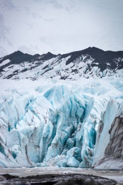 Svnafeljkull Buzulu, İzlanda 'nın manzarası. Önplanda yüksek dağlar ve kısmi donmuş buzul gölü önünde dramatik mavi buz oluşumları ve yarıklar var..