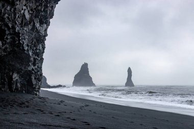 Reynisfjara 'daki bazalt mağarası. Kara kum plajı. Çarpan dalgalar ve arka planda Reynisdrangar bazalt deniz yığınları. Vik, İzlanda' da deniz ve bulutlu gökyüzüne karşı.