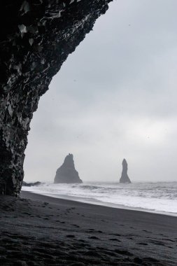 Reynisfjara 'daki bazalt mağarası. Kara kum plajı. Çarpan dalgalar ve arka planda Reynisdrangar bazalt deniz yığınları. Vik, İzlanda' da deniz ve bulutlu gökyüzüne karşı.