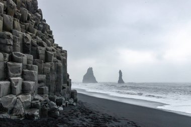 Reynisfjara 'nın kara kumsalındaki bazalt sütunlarda dalgalar ve arka planda Vik, İzlanda yakınlarında Reynisdrangar bazalt deniz yığınları var.