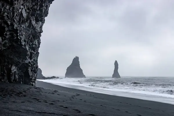 Reynisfjara 'daki bazalt mağarası. Kara kum plajı. Çarpan dalgalar ve arka planda Reynisdrangar bazalt deniz yığınları. Vik, İzlanda' da deniz ve bulutlu gökyüzüne karşı.