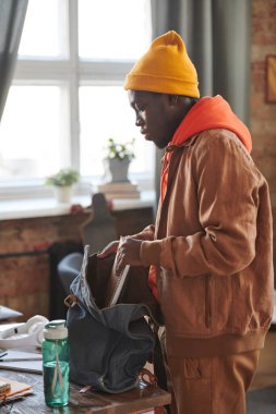 Tarz sahibi Afro-Amerikan erkek öğrenci portresi. Günlük kıyafetler giyip okula hazırlanıyor. Ders kitaplarını alıp sırt çantasına koyuyor.