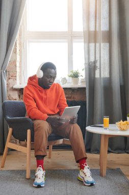 Modern genç adam gündelik kıyafetlerini giymiş sandalyede oturmuş kulaklıkla müzik dinliyor ve dijital tablette internet kullanıyor.