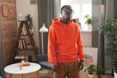 Orta boy Afro-Amerikan erkek öğrenci portresi. Turuncu kapüşonlu ve gözlüklü. Oturma odasında kameraya bakıyor.